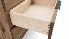 Close-up of a wooden drawer pulled open, showing a light natural wood interior and dovetail joint construction, with a dark metal pull handle on a white background.