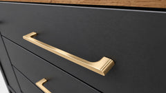 Close-up of a matte black drawer front with a brushed gold metal pull handle, showing part of a wood-grain surface above.