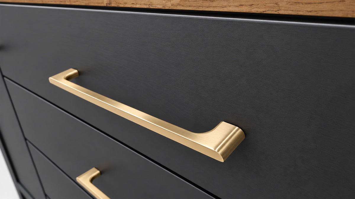 Close-up of a matte black drawer front with a brushed gold metal pull handle, showing part of a wood-grain surface above.