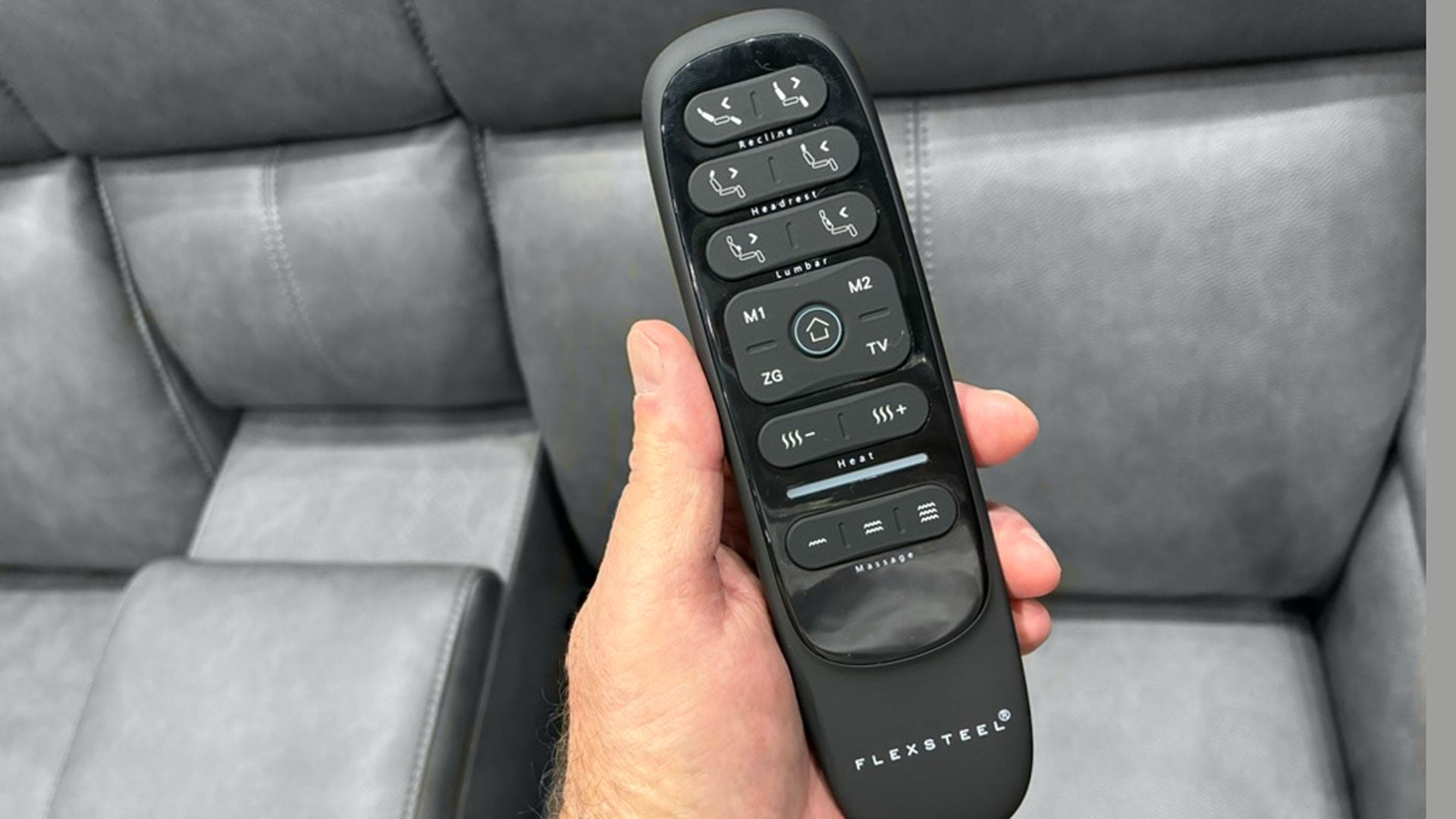 A hand holding a black FlexSteel remote control in front of a gray leather sofa.