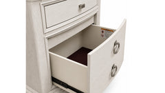 A close-up of a light gray wood nightstand or dresser drawer, with the lower drawer pulled open to show the interior and dark brown carpet lining. The drawer has a curved front and a silver metal ring pull.