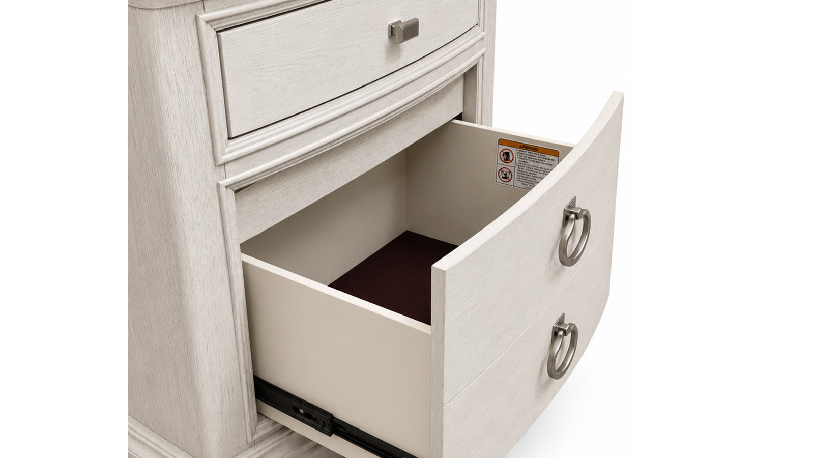 A close-up of a light gray wood nightstand or dresser drawer, with the lower drawer pulled open to show the interior and dark brown carpet lining. The drawer has a curved front and a silver metal ring pull.