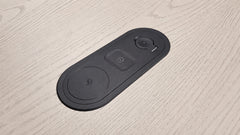 A black oval smart home control panel with three buttons, placed on a light-colored wood grain surface.