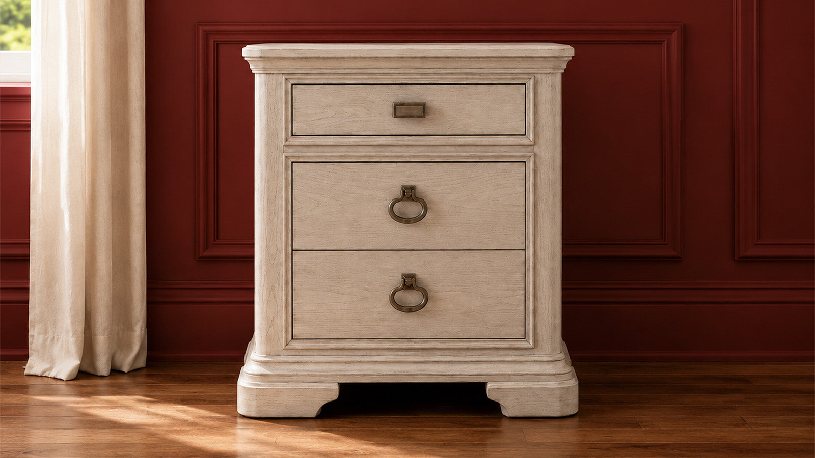 A three-drawer nightstand with a distressed cream finish and dark metal ring pulls, set against a dark red paneled wall and wooden floor.