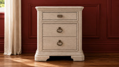 A three-drawer nightstand with a distressed cream finish and dark metal ring pulls, set against a dark red paneled wall and wooden floor.