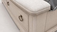 Close-up of a light beige upholstered storage bench or chest with drawers, featuring brass ring pulls and a textured fabric seat cushion.