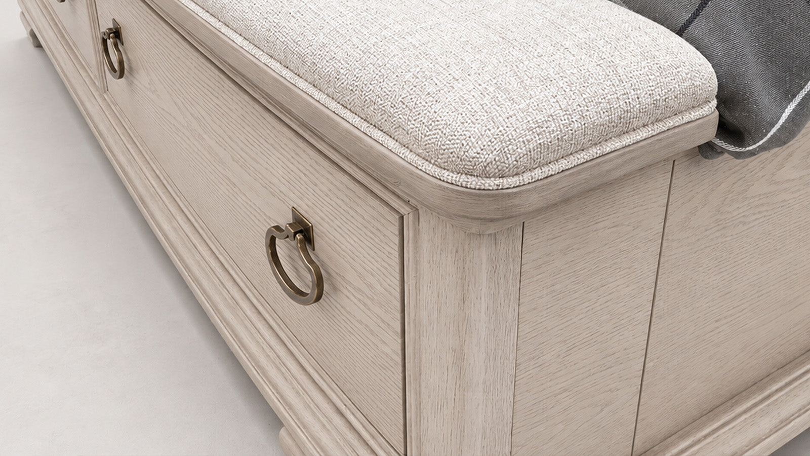 Close-up of a light beige upholstered storage bench or chest with drawers, featuring brass ring pulls and a textured fabric seat cushion.