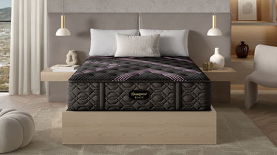 A king size Beautyrest Black Series 2 Firm Tight Top Mattress displayed on a bed frame in a modern bedroom setting with a decorative bedskirt, bed pillows, and a nightstand with a lamp.