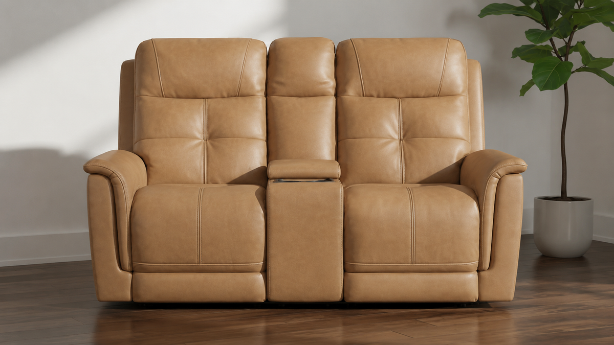 A tan leather reclining loveseat with two seatbacks, wide armrests, and a center console between the seats, shown in a staged room with wood flooring and a potted plant.