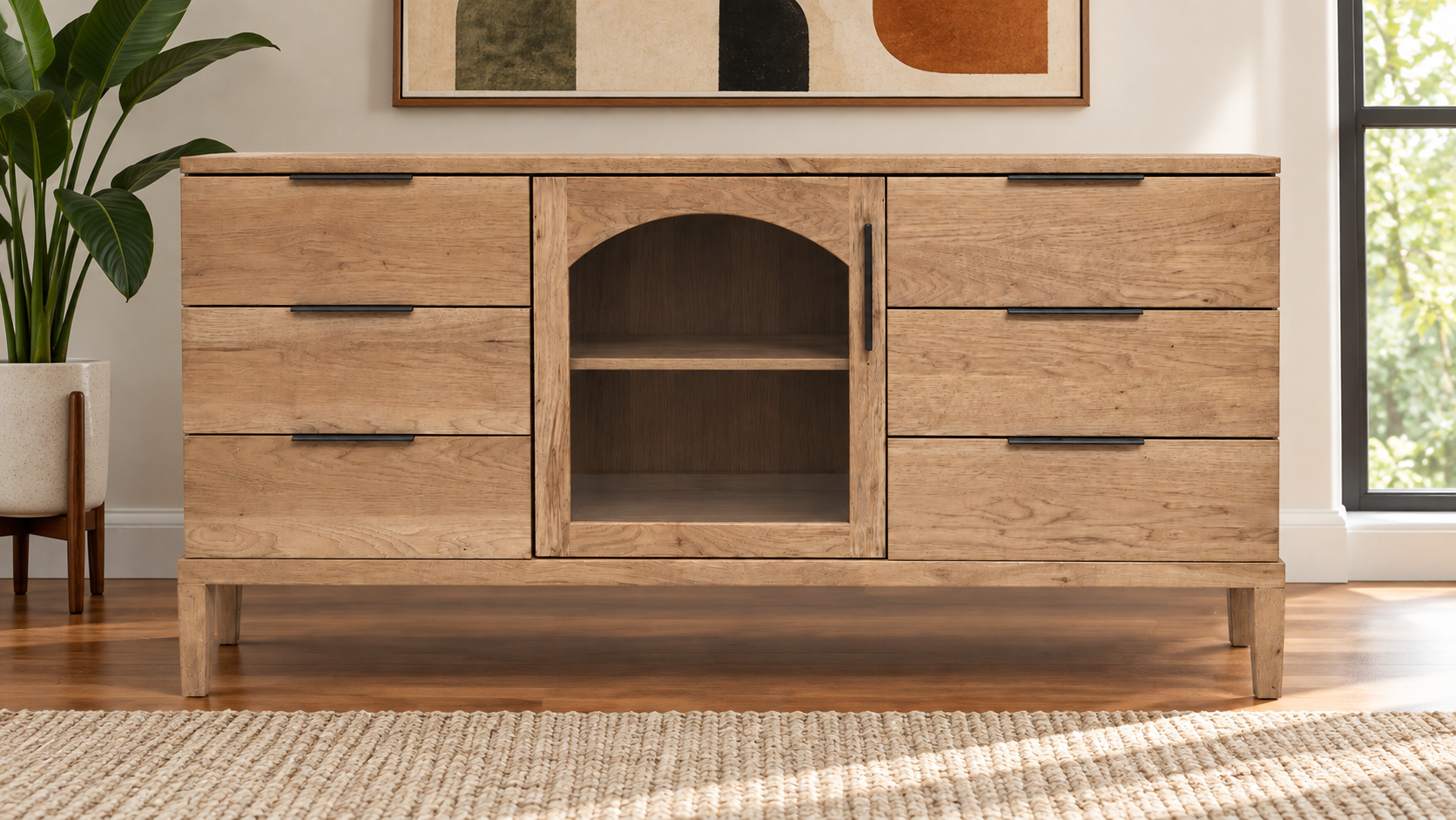 A light oak wood sideboard cabinet with five drawers, a central arched open shelf, and dark metal handles, shown in a styled room setting with a plant, wall art, and window.