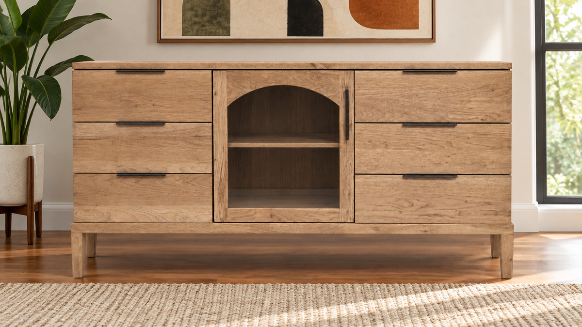 A light oak wood sideboard cabinet with five drawers, a central arched open shelf, and dark metal handles, shown in a styled room setting with a plant, wall art, and window.