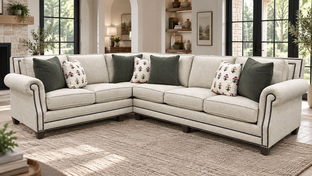 An L-shaped sectional sofa upholstered in light beige fabric with dark nailhead trim, styled in a bright living room with dark green and floral accent pillows.