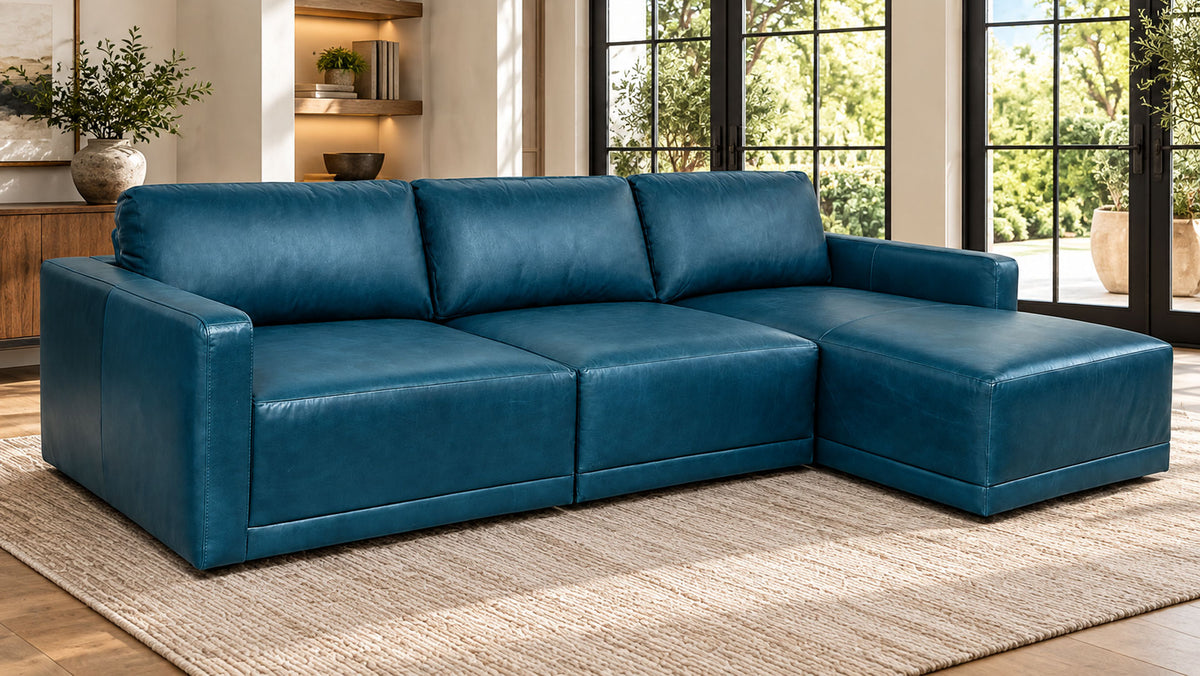 A modern sectional sofa with a right-facing chaise lounge in deep teal blue leather, styled on a beige woven rug in a bright living room.