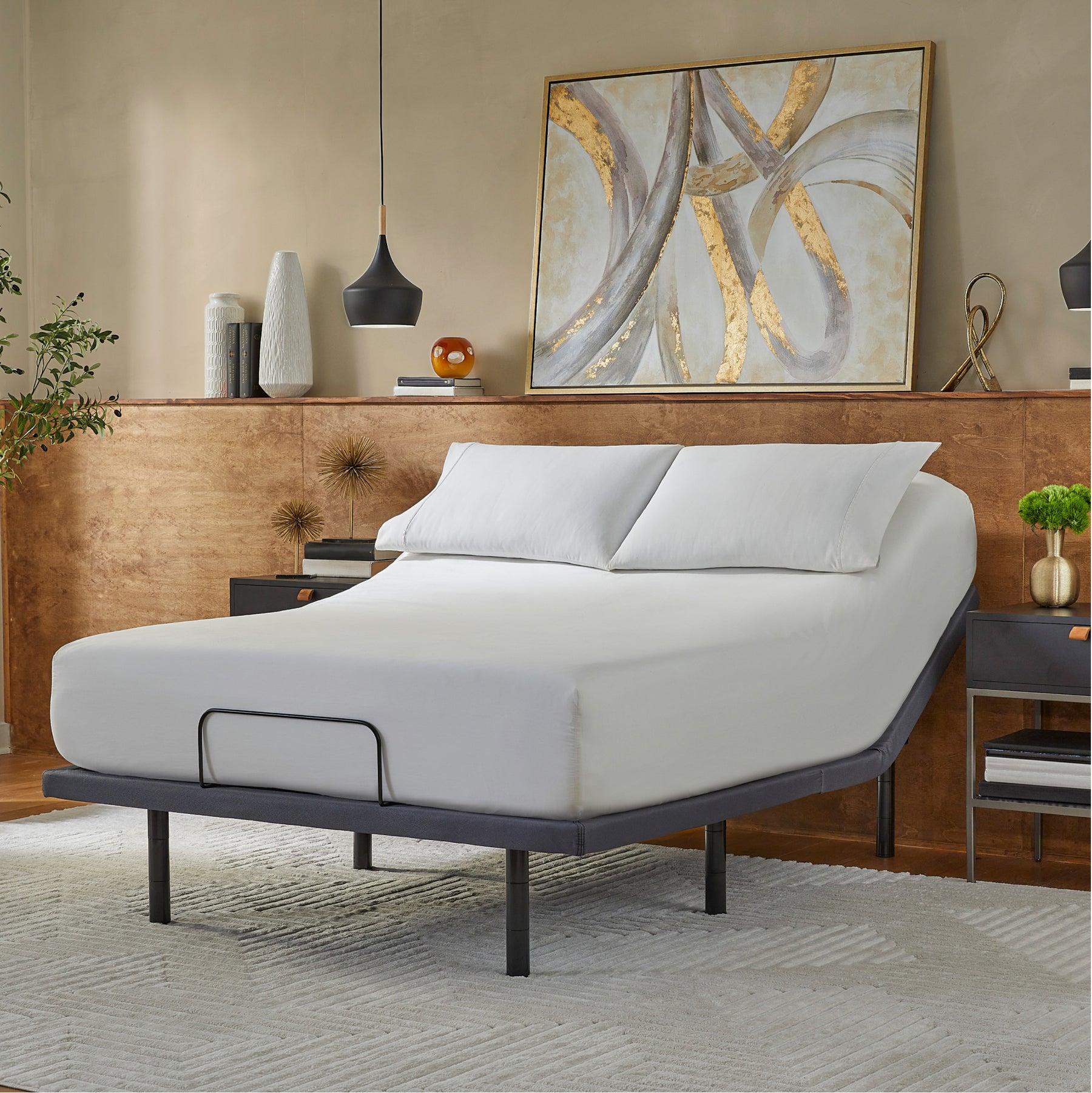 An adjustable metal base of a bed frame with a mattress on it, positioned in a room with minimalist decor. The mattress is accompanied by two pillows, and the room has a large painting on the wall, a bedside lamp, and a small plant.
