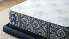 Un colchón Serta visto desde un ángulo, apoyado sobre una base de cama oscura sobre una alfombra de color beige claro. El colchón presenta una superficie superior blanca con textura y un panel lateral estampado en azul marino, gris y blanco, en el que se aprecia el logotipo de Serta.