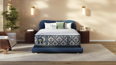 Una cama de lujo con cabecero y piecero tapizados en azul marino, que cuenta con un colchón estampado con motivos geométricos en azul, blanco y gris. La cama está decorada con cojines blancos y verdes en un dormitorio acogedor y moderno, con una pared beige, apliques de pared y una alfombra de tonos claros.