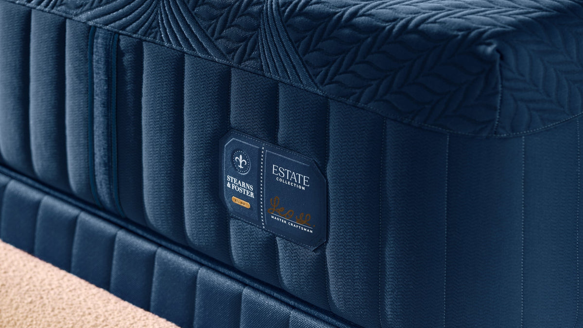 A close-up of a dark blue upholstered mattress with quilted top and side panels, showing a brand label that reads Estate Textiles and Serta, set against a neutral beige carpet background.