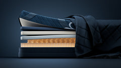 A close-up side view of a mattress stack with layered bedding, including a navy quilted blanket, patterned blue sheets, white and gray mattress layers, and a warm tan inner layer, set against a dark navy studio background.