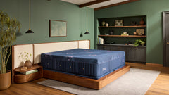 A blue mattress on a wooden platform bed in a styled bedroom with green walls, wood ceiling beams, pendant lights, a rug, and built-in shelving.