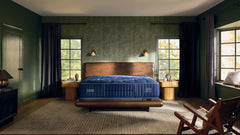 A luxury bedroom scene featuring a dark blue tufted mattress on a wooden bed frame, set in a styled room with dark green walls, textured wallpaper, and a wooden rocking chair in the foreground.