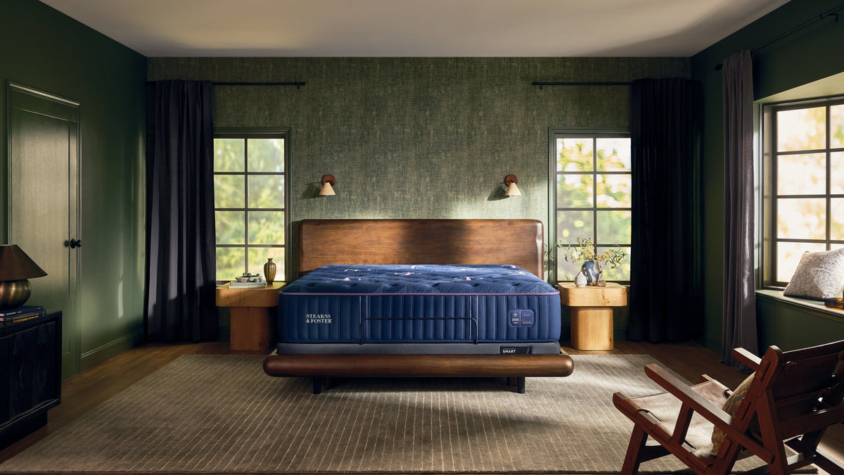 A luxury bedroom scene featuring a dark blue tufted mattress on a wooden bed frame, set in a styled room with dark green walls, textured wallpaper, and a wooden rocking chair in the foreground.
