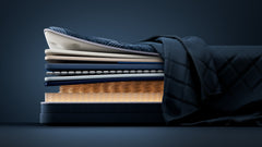 A stack of layered mattress toppers or bedding components in dark navy, cream, and blue tones, with a quilted navy fabric draped over the stack, set against a dark navy studio background.