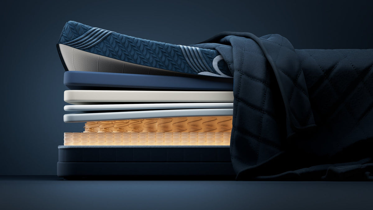 A stack of layered bedding or mattress toppers in dark navy, blue, white, and tan, with a dark quilted fabric draped over the top, photographed against a dark blue studio background.