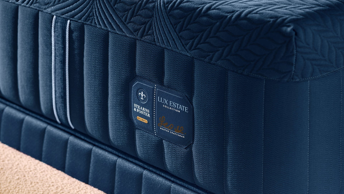 A close-up of a dark navy blue mattress with quilted side panels and a visible Lux Estate brand label, set against a neutral beige background.