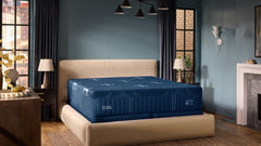 A luxury bed with a deep blue mattress and a beige upholstered platform headboard and footboard, set in a modern bedroom with blue walls, dark curtains, and wood floors.