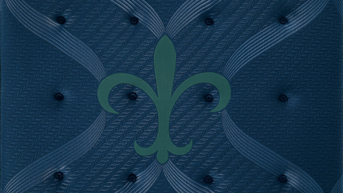 A square tufted mattress topper or mattress top in dark navy blue, featuring a subtle textured herringbone pattern, tufted button details, and a central green fleur-de-lis logo.