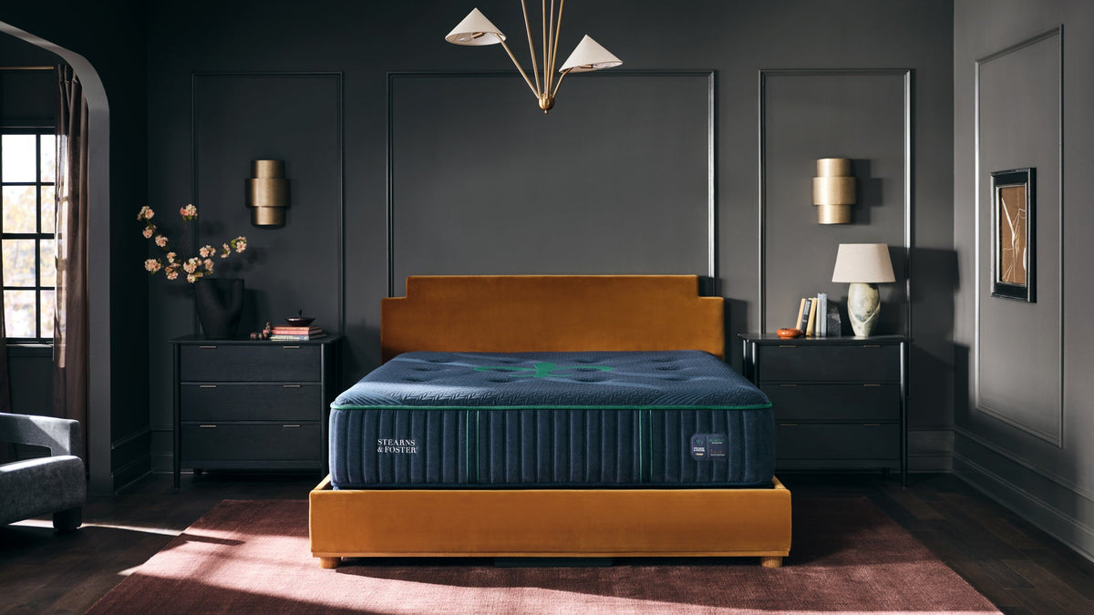Un colchón de lujo expuesto en un dormitorio decorado. El colchón tiene la parte superior de color azul oscuro y el lateral acanalado de color azul marino con un logotipo verde, y descansa sobre un somier de color amarillo mostaza.