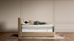 A person lies on their back on a mattress on a bed frame in a styled bedroom setting with beige paneled walls and a wood floor.