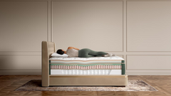 A person lies on their back on a high-profile mattress on a platform bed frame, set against a beige paneled wall and wood floor.