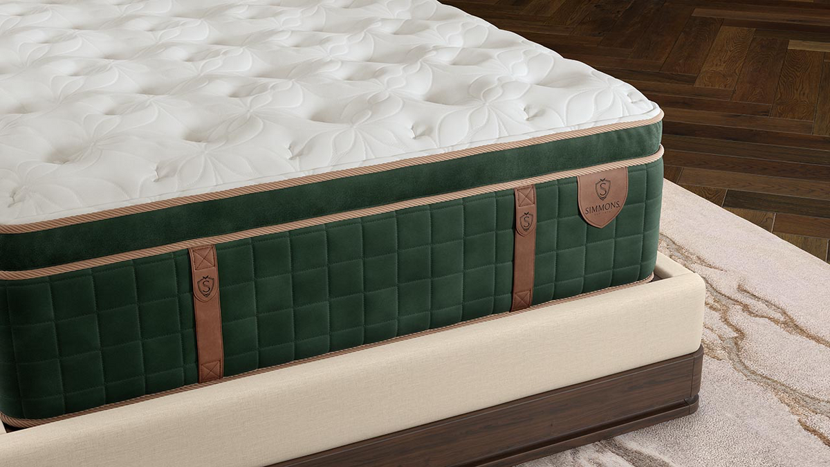 A close-up of a high-end mattress showing its white tufted top, dark green quilted side panels, and brown leather straps, resting on a matching upholstered bed frame in a bedroom setting.