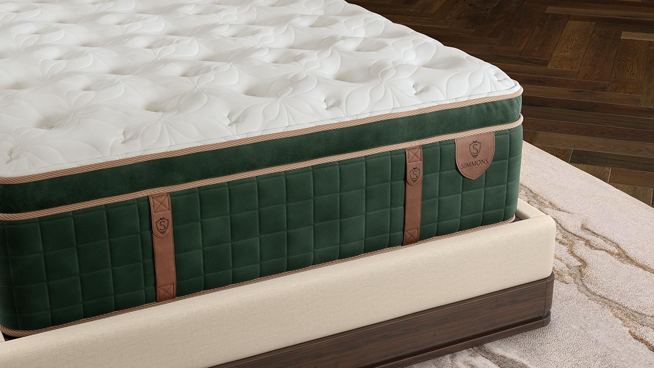 A close-up of a high-end mattress showing its white tufted top, dark green quilted side panels, and brown leather straps, resting on a matching upholstered bed frame in a bedroom setting.