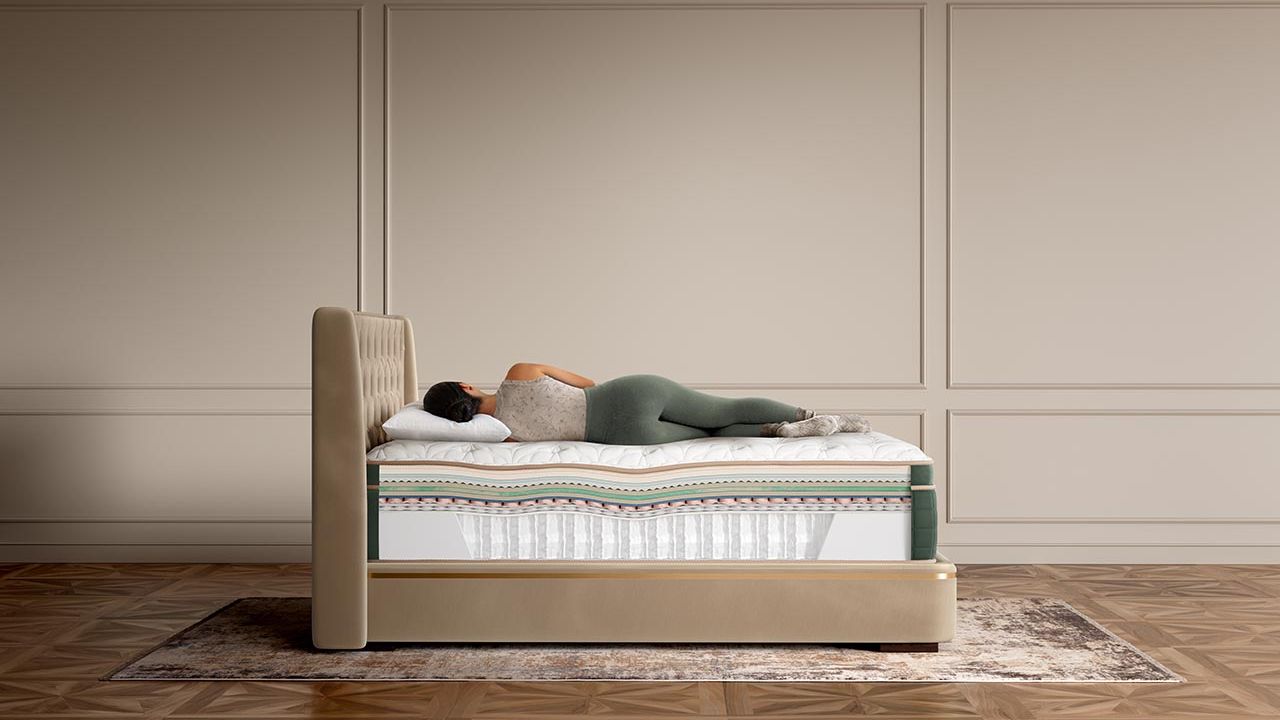 A person lies on their back on a mattress in a bed with a beige upholstered frame, set against a beige paneled wall and wood floor.