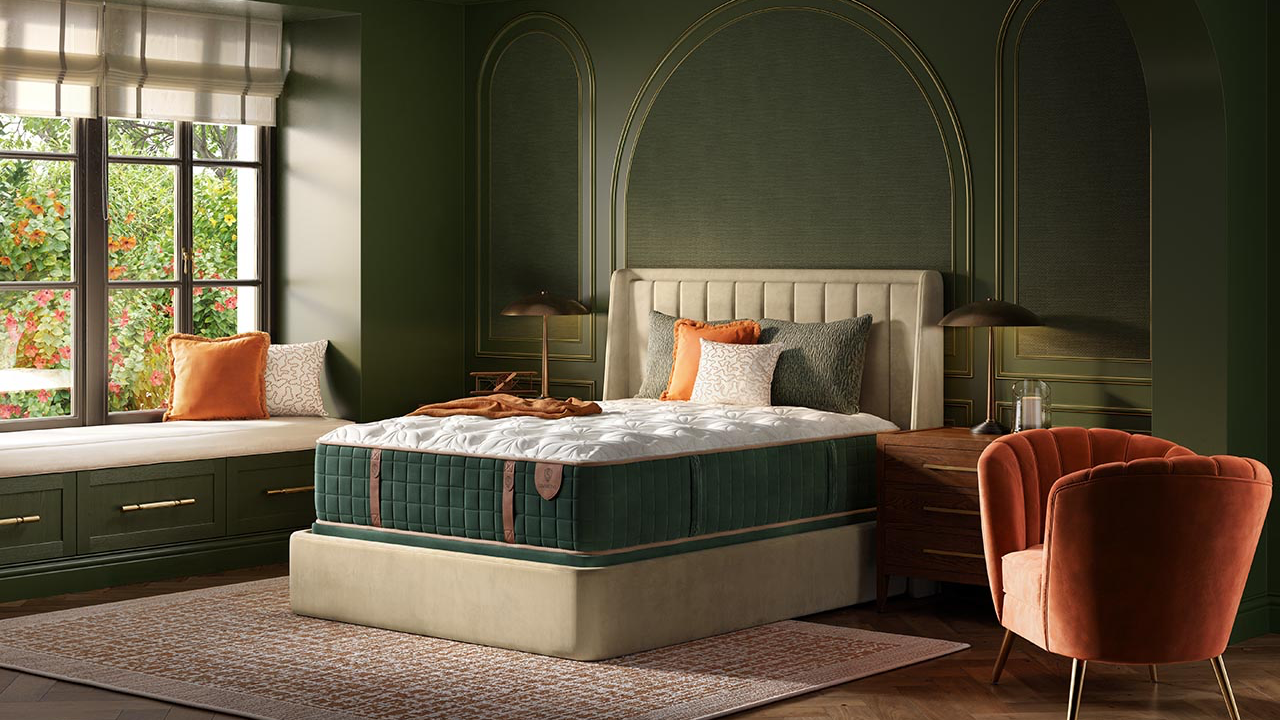 Una cama de lujo con cabecero capitoné en color crema y colchón en verde intenso, situada en un dormitorio decorado en verde oscuro con un sillón naranja, un asiento junto a la ventana y una alfombra estampada.