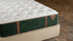 Un colchón de lujo fotografiado en ángulo en un dormitorio, con una parte superior capitoné blanca, un panel lateral acolchado de color verde oscuro con correas de imitación de cuero de color beige claro y una base de color beige, sobre un suelo de parqué de madera y una alfombra estampada.