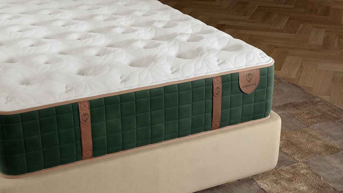 Un colchón de lujo fotografiado en ángulo en un dormitorio, con una parte superior capitoné blanca, un panel lateral acolchado de color verde oscuro con correas de imitación de cuero de color beige claro y una base de color beige, sobre un suelo de parqué de madera y una alfombra estampada.