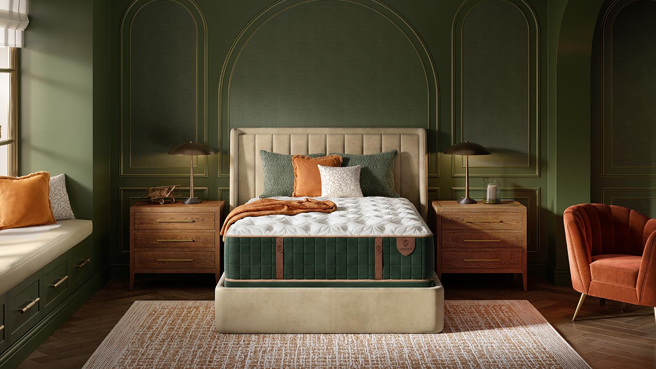Una cama de lujo con un cabecero acolchado de color crema y un colchón capitoné verde, situada en un dormitorio verde ricamente decorado con mesitas de noche de madera, lámparas, un sillón naranja y una alfombra estampada.