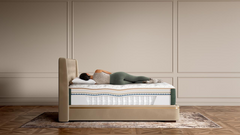 A person lies on their back on a mattress in a bed with a beige upholstered frame, set against a beige paneled wall and wood floor.