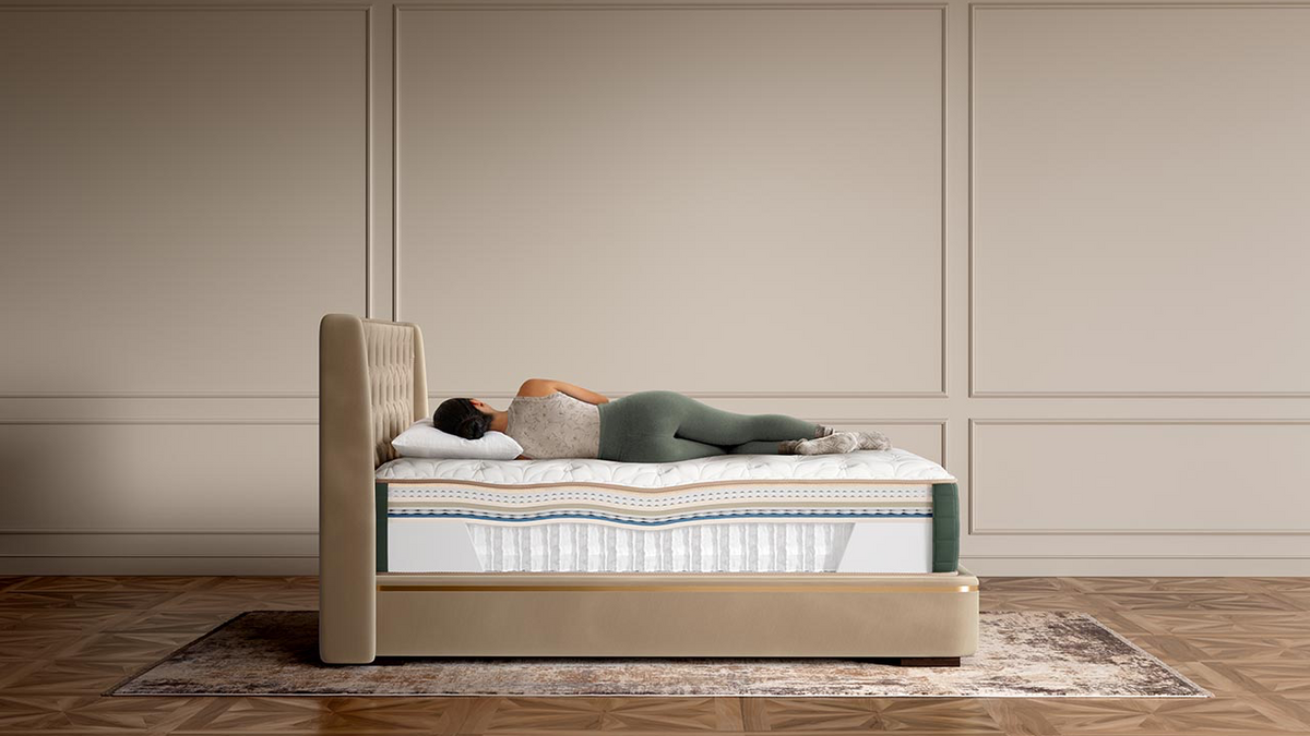 A person lies on their back on a mattress in a bed with a beige upholstered frame, set against a beige paneled wall and wood floor.