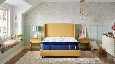 A queen-sized Stearns & Foster mattress with a pillowtop design, displayed in a bedroom setting. The mattress is set in a room with light green walls, a ceiling fan, and is dressed with a blue and white bedding set.