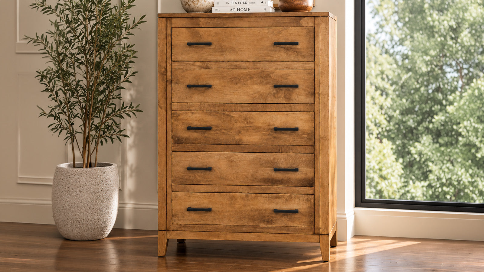 A five-drawer wooden chest of drawers with a warm medium brown finish and black metal handles, positioned in a room next to a window and a potted plant.