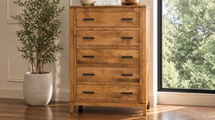 A five-drawer wooden chest of drawers with a warm medium brown finish and black metal handles, positioned in a room next to a window and a potted plant.