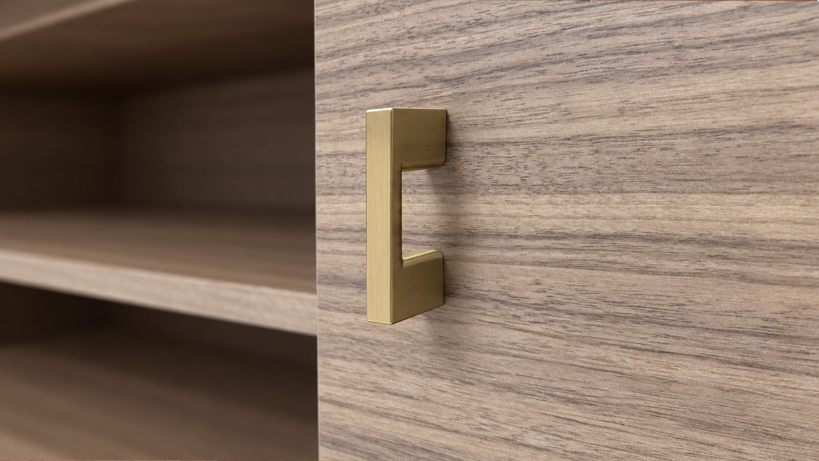A modern, minimalist L-shaped cabinet pull handle in a brushed brass/gold finish, mounted on a wood-grain cabinet door next to open shelves.