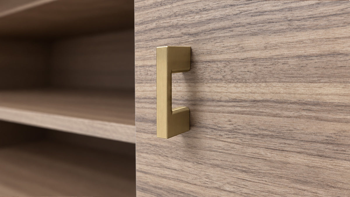 A modern, minimalist L-shaped cabinet pull handle in a brushed brass/gold finish, mounted on a wood-grain cabinet door next to open shelves.