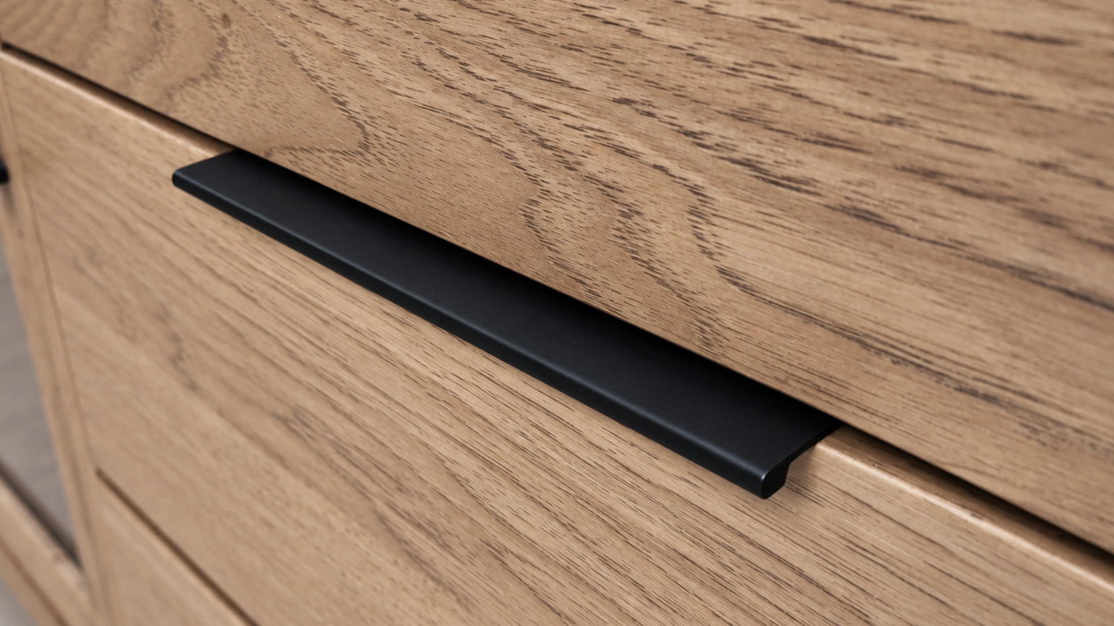 A close-up of a modern matte black drawer pull handle mounted on a light oak wood cabinet.