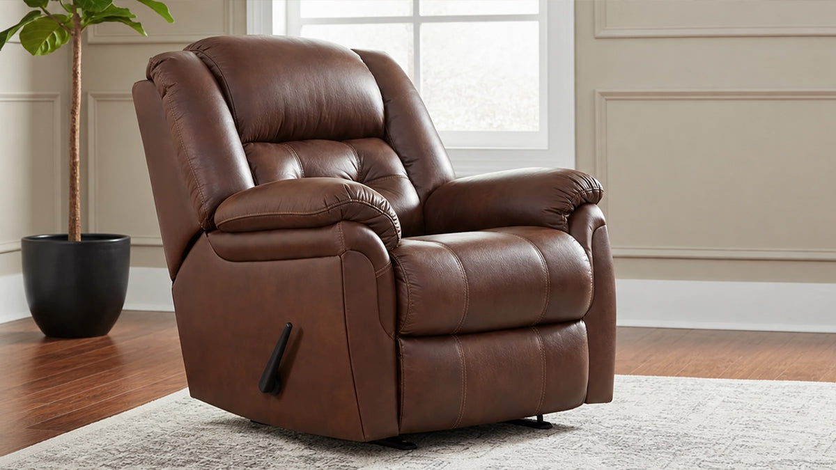 A brown micro recliner chair with a tufted backrest and padded armrests, shown in a staged living room setting with a window, wood floor, rug, and potted plant.