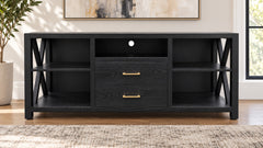 A black wood media console with two drawers featuring gold handles, open side shelves, and X-shaped metal support legs, shown in a styled living room setting.